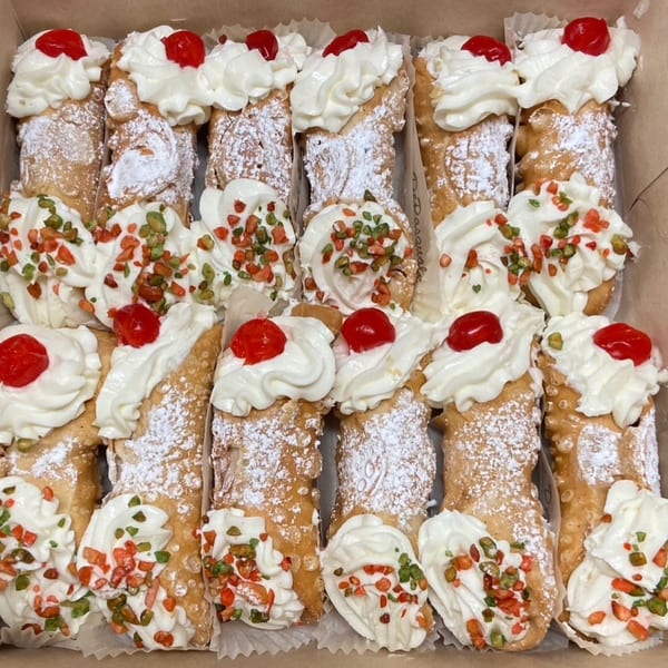 A box filled with cannolis, some topped with cherries and others with green and red sprinkles.