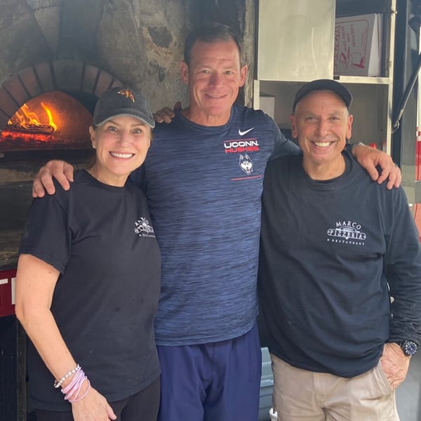 Three smiling people, two wearing Marco Pizzeria shirts, stand in front of a wood-fired pizza oven.