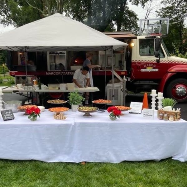 Marco Pizzeria food truck and tent with a table of pizzas and red roses on a lawn.
