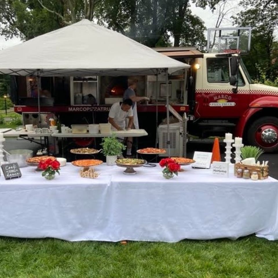 Marco Pizzeria food truck and tent with a table of pizzas and red roses on a lawn.