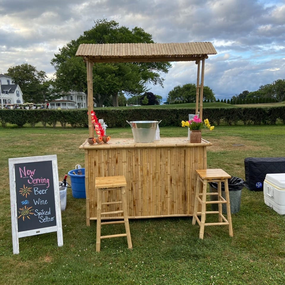 A bamboo bar with two stools, a "Now Serving" sign, and coolers on a grassy lawn.