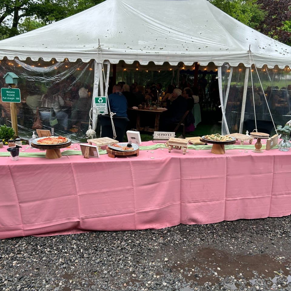 A pink-covered buffet table with pizzas and other dishes outside a white tent with diners.