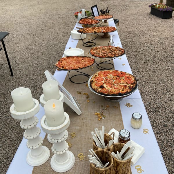 A long outdoor table laden with various Marco Pizzeria pizzas, candles, and serving supplies.