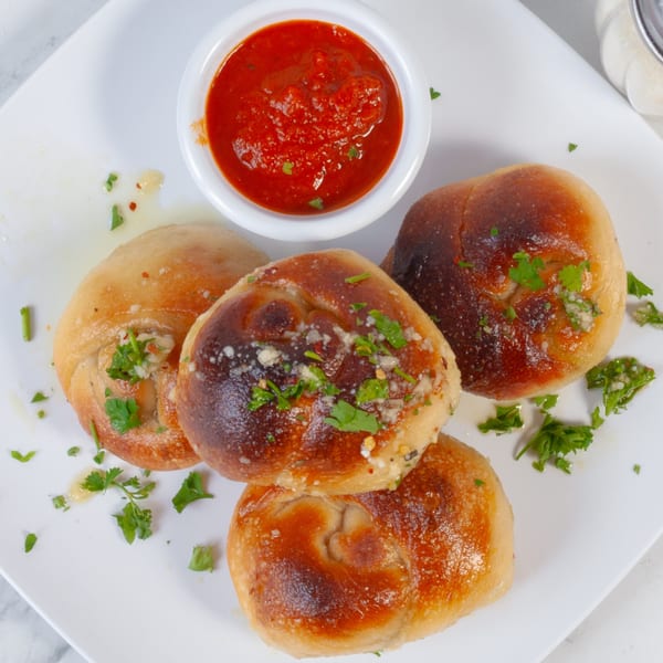 Four golden-brown garlic knots with parsley and cheese, served with a side of red marinara sauce.
