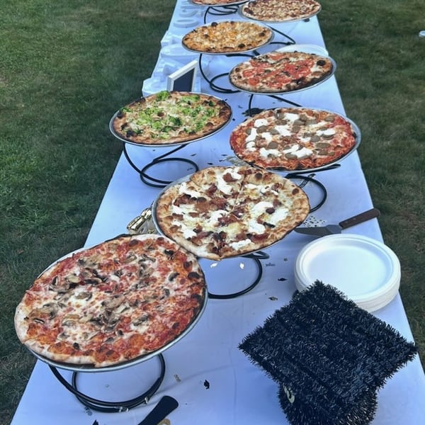 A long table with a white tablecloth holds several Marco Pizzeria pizzas on tiered stands outdoors.