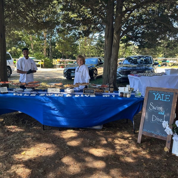 Two Marco Pizzeria chefs stand behind a blue table laden with pizzas and Italian food outdoors.