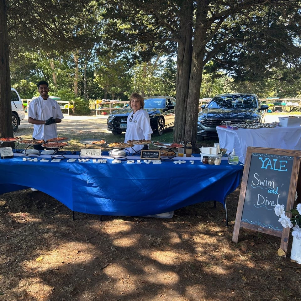 Two Marco Pizzeria chefs stand behind a blue table laden with pizzas and Italian food outdoors.