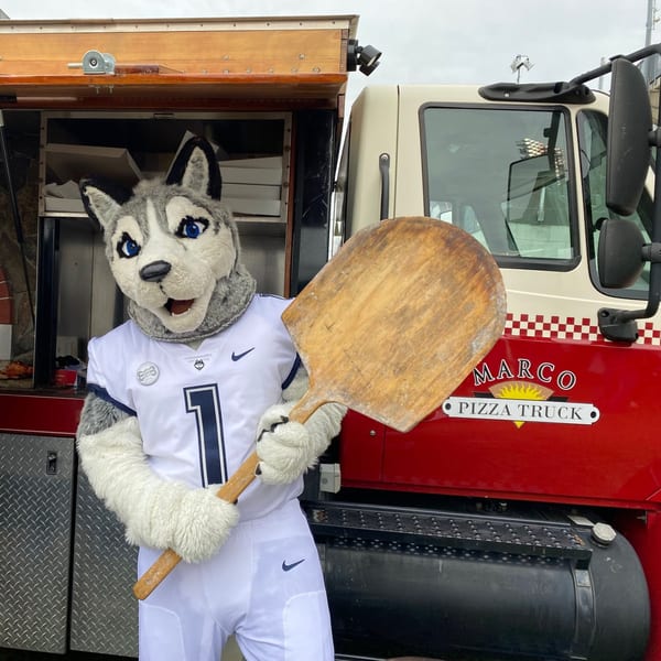 UConn husky mascot holding a pizza peel in front of the Marco Pizza Truck.