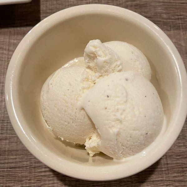 Three scoops of creamy vanilla bean gelato in a white bowl on a wooden table.