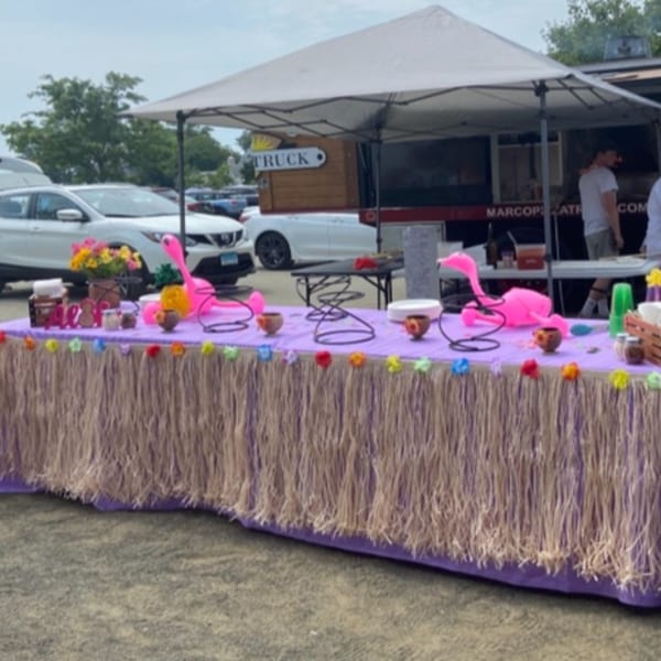 A purple table with a grass skirt, pink flamingos, and flowers, with a food truck in the background.
