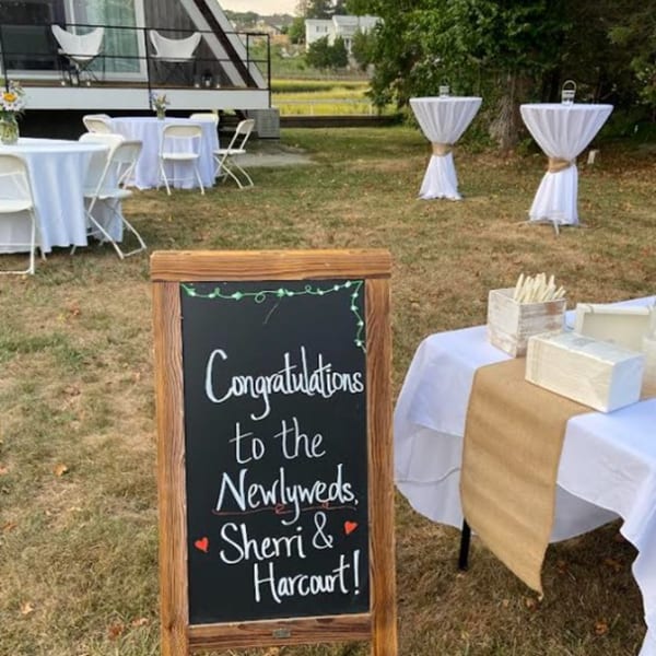 Outdoor wedding reception setup with a chalkboard sign, white tables, and cocktail tables on a grassy lawn.