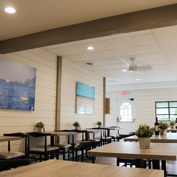 Bright, empty dining room at Marco Pizzeria with light wood tables, dark chairs, and nautical wall art.
