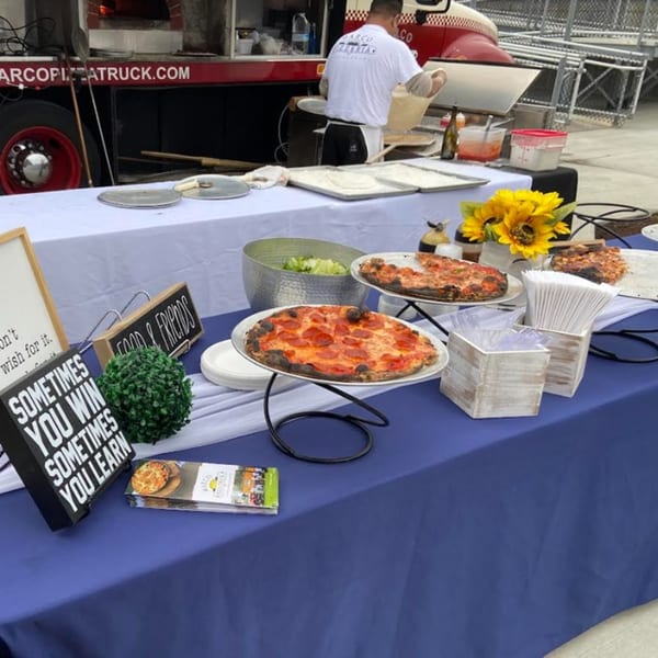 Marco Pizzeria food truck serving fresh pizzas and salad on a blue and white draped table.