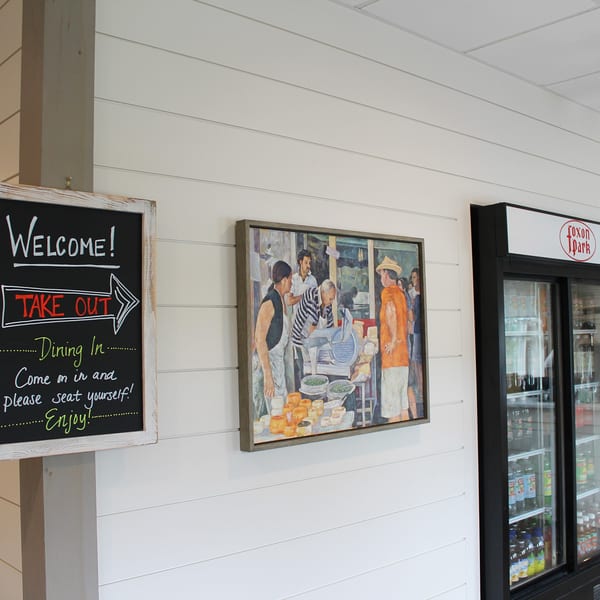 Chalkboard sign with "Welcome! Take Out Dining In" next to a painting and a beverage cooler.