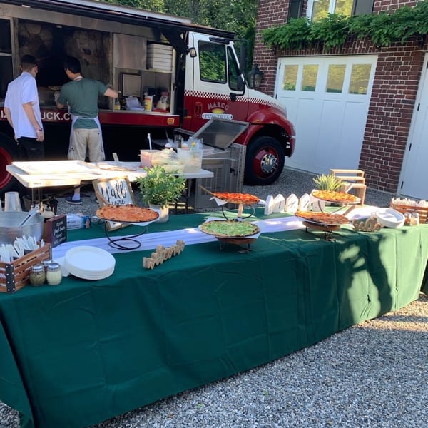 Marco Pizzeria food truck with chefs and a buffet table laden with pizzas and serving supplies.