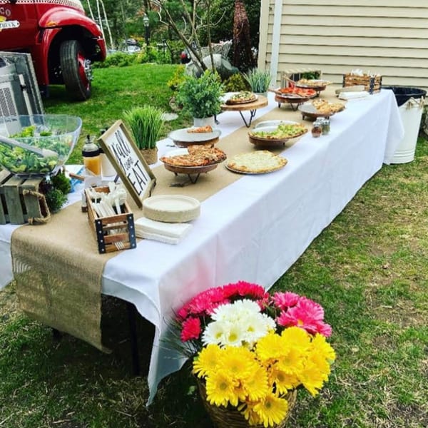 Outdoor catering table with Marco Pizzeria's pizzas, salad, and a red food truck in the background.