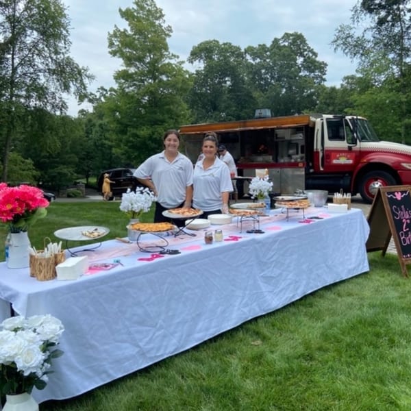 Two women stand behind a table laden with pizzas and flowers, with a Marco Pizzeria food truck behind them.