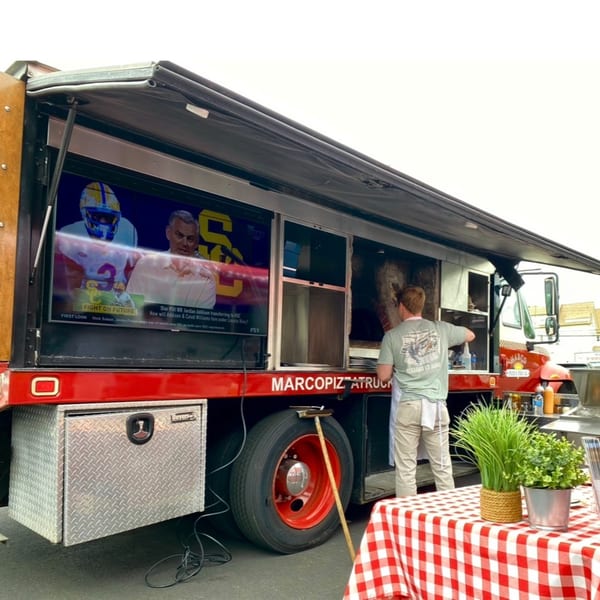 A Marco Pizzeria food truck with a TV screen showing sports, and a chef working inside.