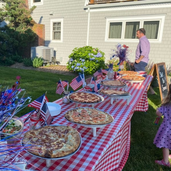 Outdoor party with pizzas on a red and white checkered table, American flags, and people.
