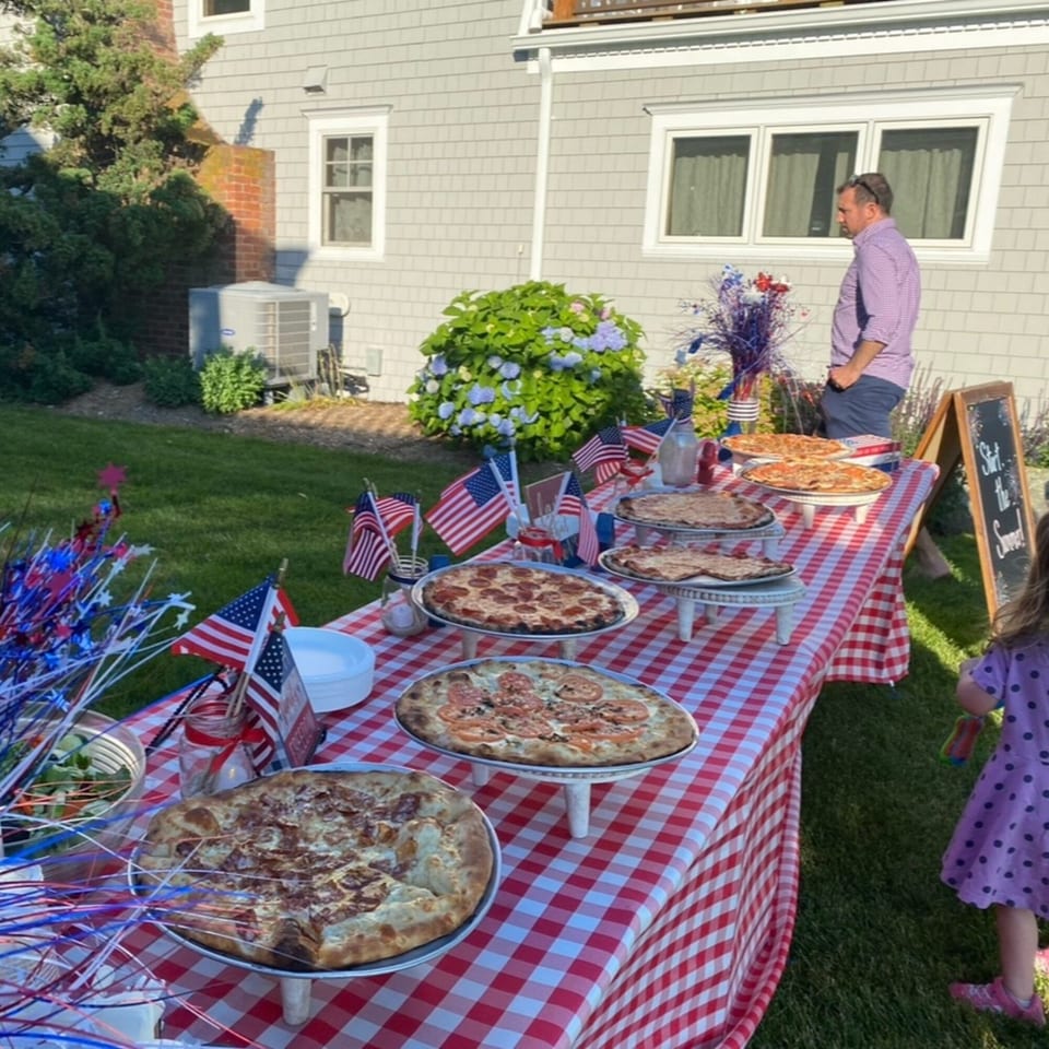 Outdoor party with pizzas on a red and white checkered table, American flags, and people.