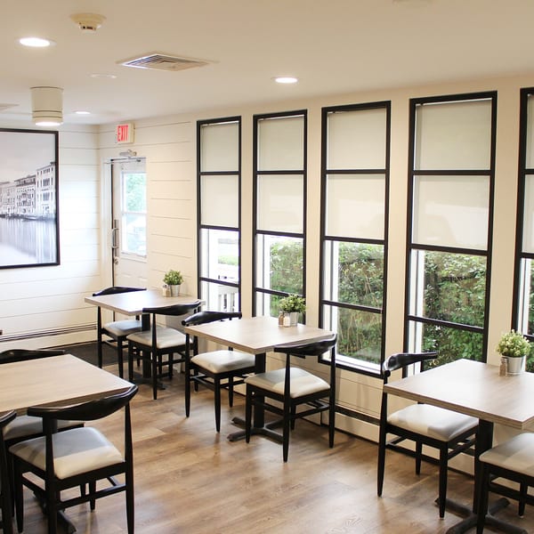 Bright, empty dining room at Marco Pizzeria with light wood tables, black chairs, and large windows.