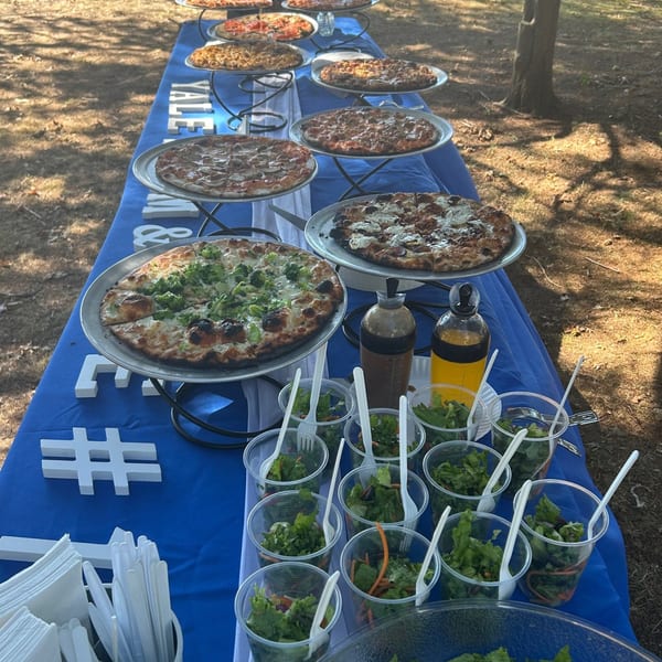 A long table covered in a blue cloth, laden with multiple pizzas and individual salad cups outdoors.