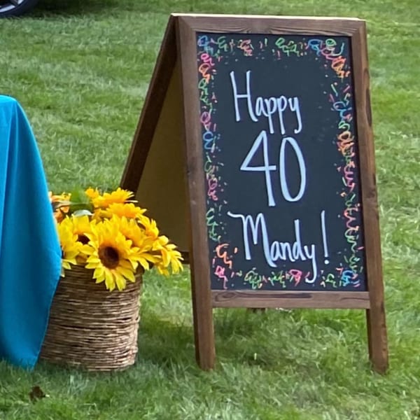 A chalkboard sign on grass reads "Happy 40 Mandy!" next to a basket of sunflowers.
